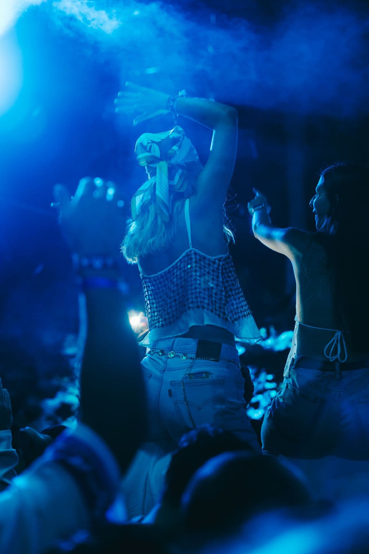 A dancer in a mesh top with both arms above her head, lit deep blue through the haze.