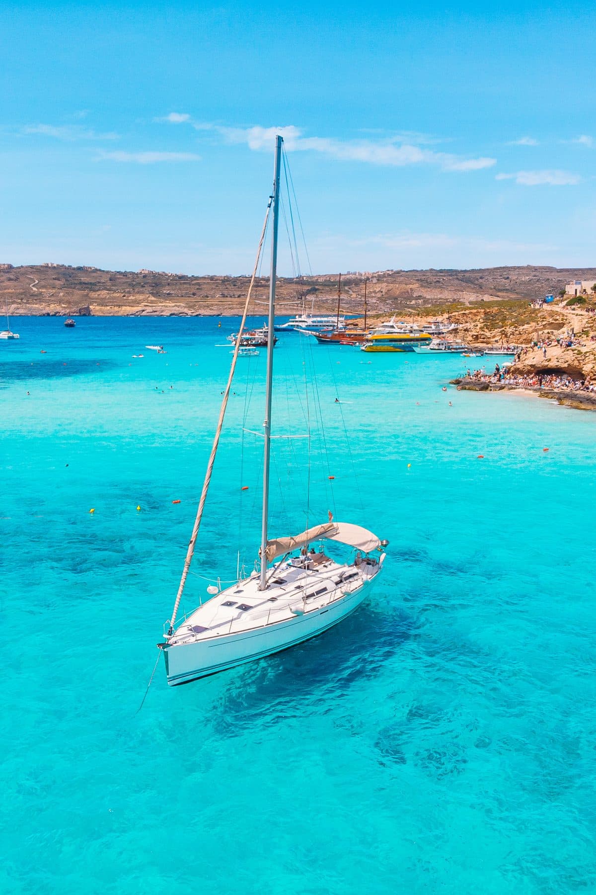A sailboat anchored in the turquoise shallows of the Blue Lagoon at Comino, boats and swimmers along the rocky shore behind it.