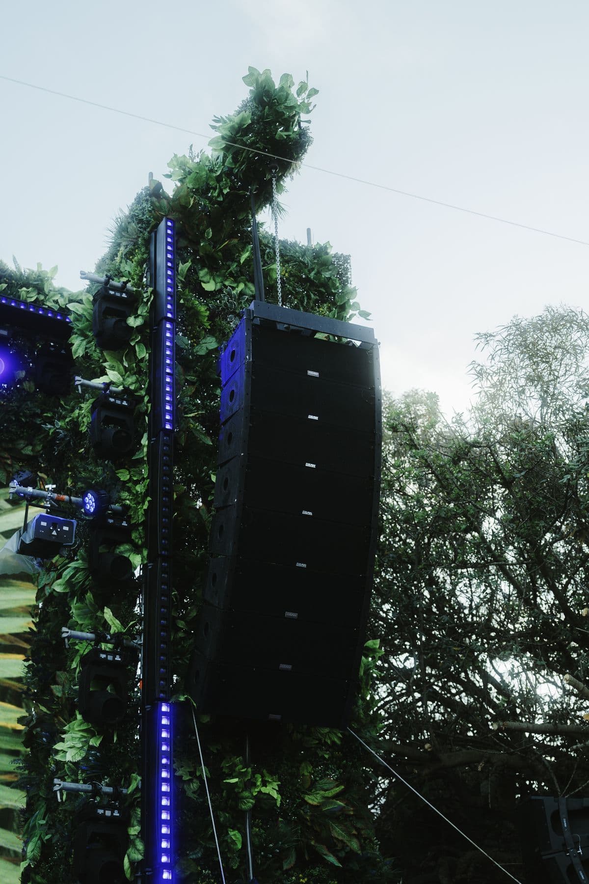 A hanging line-array speaker stack framed by foliage and blue uplighting, in daylight.